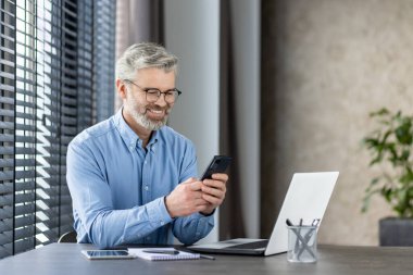 Ofis masasında dizüstü bilgisayar ve defterle otururken akıllı telefon kullanarak gülümseyen olgun bir iş adamı. Mutlu ve üretken görünüyor..