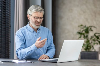 Üst düzey işadamı, modern ofis ortamında dizüstü bilgisayar ve kulaklık kullanarak bir video görüşmesi yapıyor..