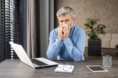 İş adamı, evinde bilgisayarlı, peçeteli, defterli ve gözlüklü bir masada çalışırken kendini iyi hissetmiyor..