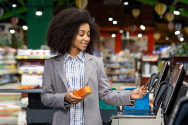Gri ceketli, gülümseyen bir kadın bir markette kendini kontrol eden bir büfe kullanıyor. Makineyle etkileşime girerken elinde turuncu bir ürün tutuyor..