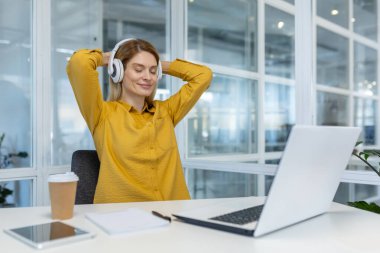 Kulaklıkla rahatlayan ve modern bir ofiste masasında müzik dinlemekten zevk alan bir kadın. Sakin ve odaklanmış görünüyor..
