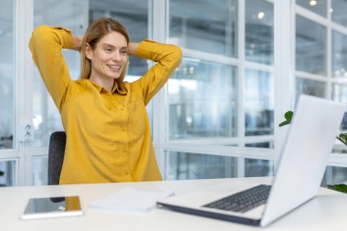 Sarı tişörtlü gülümseyen kadın, bir ofis masasında dizüstü bilgisayarla otururken dinleniyor..
