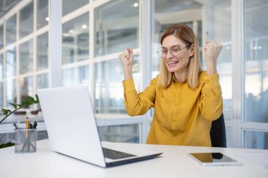 Sarı gömlekli iş kadını ofis masasında başarısını dizüstü bilgisayarla kutluyor, sevinç ve heyecan ifade ediyor..