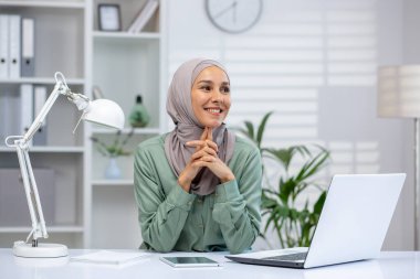 Başörtüsü takan ve masasında dizüstü bilgisayarla çalışan gülümseyen genç bir kadın. Parlak, profesyonel bir atmosferi olan modern ofis..