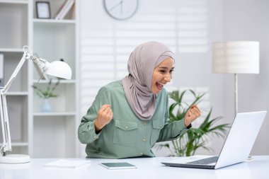 Ev ofisinde başarısını kutlayan tesettürlü neşeli bir kadın elinde dizüstü bilgisayar ve tabletle bir masada oturuyor..