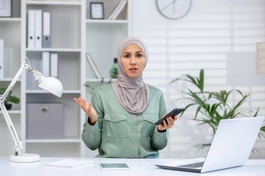 Ev ofisinden çalışan tesettürlü bir kadın, elinde akıllı telefon ve kafa karışıklığı içinde el kol geziyor. Dizüstü bilgisayarlı ve belgeli modern çalışma alanı.