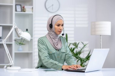 Ev bilgisayarında uzaktan çalışan, modern ve parlak bir ofis ortamında kulaklık takan tesettürlü bir kadın..