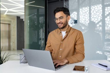 Hintli genç adam modern bir ofiste çalışıyor, bir masada oturuyor, gülümsüyor ve konsantre bir şekilde klavyeye yazıyor..