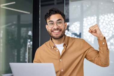 Ofisteki genç bir Müslüman adamın yakın plan fotoğrafı ve kameraya eliyle bir zafer jesti gösteriyor. Başarı ve başarıdan etkilenmiş, gülümseyerek kameraya bakıyor..
