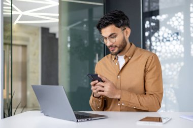 Modern ofiste dizüstü bilgisayarla çalışırken genç bir profesyonel telefonu kontrol ediyor. İş ortamı, günlük kıyafetler, modern teknoloji..