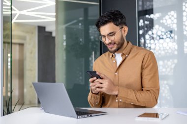 Kahverengi gömlekli genç bir Hintli ofiste oturuyor ve cep telefonu kullanıyor, mesajlar yazıyor, mesajlar yazıyor..