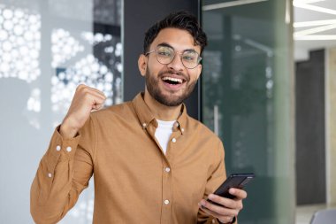 Gözlüklü genç adam modern bir iç mekanda akıllı telefon tutarken tezahürat ve kutlama yapıyor. Başarı ve mutluluk kavramı.