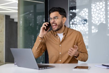 Hüsrana uğramış işadamı, modern ofis ortamında masasında çalışırken yoğun bir telefon görüşmesi yapıyor..