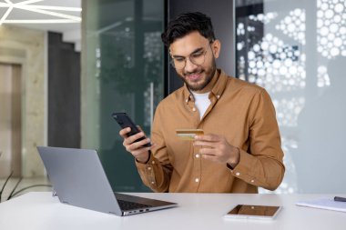 Gülümseyen genç Müslüman iş adamı masadaki ofiste oturuyor, telefonu elinde tutuyor ve bir kredi kartı kullanıyor, internet hesabı..