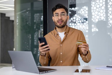 Endişeli genç Hintli bir adam ofiste dizüstü bilgisayarla oturuyor, elinde kredi kartı ve telefon var, şok içinde kameraya bakıyor..