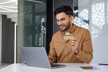 Genç Hintli bir adam bir ofiste oturuyor, bir dizüstü bilgisayar üzerinde çalışıyor ve bir kredi kartı kullanıyor..