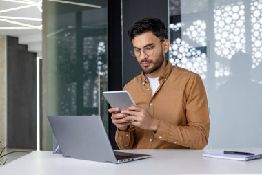 Modern bir ofis ortamında çalışırken tablet kullanarak profesyonelce odaklanmış. Adam meşgul ve konsantre olmuş gibi görünüyor..
