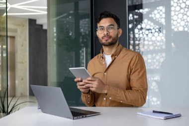 Genç Müslüman bir adamın portresi, elinde bir bilgisayarla büroda oturuyor, elinde bir tablet ve dikkatlice kameraya bakıyor..