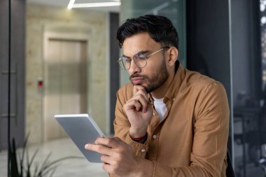 Modern bir ofis ortamında dijital tablet kullanan sıradan giysili bir adam. Çalışan ya da okuyan kişi.