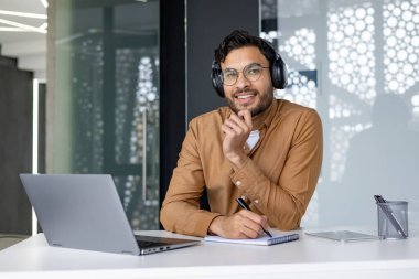 Modern ofis ortamında dizüstü bilgisayarıyla çalışan kulaklık takan gülümseyen bir adam. Not alırken meşgul ve üretken görünüyor..