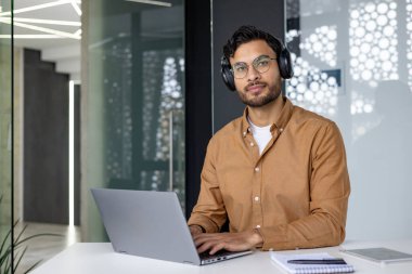 Ofiste oturan, kulaklık takan, dizüstü bilgisayarda çalışan ve kameraya ciddi ciddi bakan Hintli bir adamın portresi..