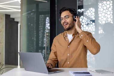 .Ofiste dizüstü bilgisayarla çalışan, kulaklıkla masada oturan ve müzik dinleyen, dinlenen, şarkı söyleyen Hintli genç adam..