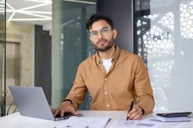 Ofiste dizüstü bilgisayar ve belgelerle çalışan, elinde kalem tutan ve kameraya bakan Hintli genç bir adamın portresi..