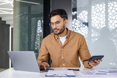 Ofiste çalışan ciddi ve odaklanmış genç Hintli bir adam elinde hesap makinesiyle veri ve muhasebe ile çalışıyor..
