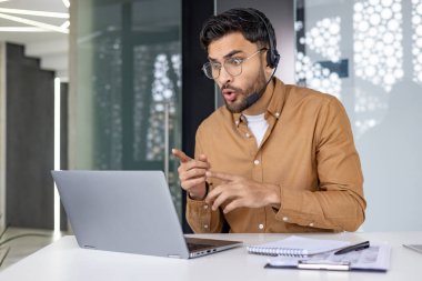 Ciddi bir Hintli genç adam ofiste kulaklıkla oturuyor ve dizüstü bilgisayara bakıyor, haberleri okuyor, bir video görüşmesinde konuşuyor..