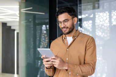 Genç gülümseyen Müslüman erkek programcı ve tasarımcı modern ofiste duruyor ve tablet kullanıyor..