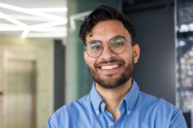 Modern bir ofiste gülümseyen ve kameraya bakan gözlüklü ve mavi gömlekli genç bir Hintlinin yakın plan portresi..