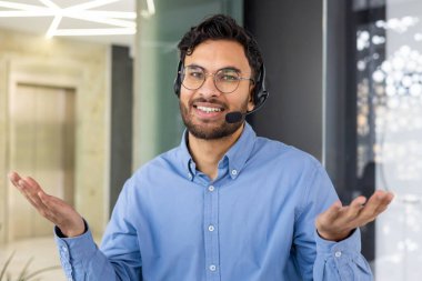Gözlüklü ve mavi gömlekli gülümseyen Hintli bir adamın yakın plan portresi ofiste, kameraya bakıyor ve ellerini açıyor..