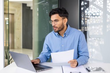 Dizüstü bilgisayarda çalışan ve modern ofis ortamında belgeleri inceleyen gözlüklü genç bir profesyonel. Konsantrasyon ve verimlilik kavramı.