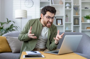 Gözlüklü ve yeşil tişörtlü kızgın genç adam evdeki kanepede oturuyor ve ekranda duygusal olarak jest yapıyor, kötü haberler alıyor, video konuşmasında konuşuyor..