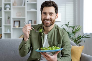 Sağlıklı bir hayat süren, kanepede oturan, tabaktan taze salata yiyen ve kameraya bakan gülümseyen genç bir adamın yakın plan fotoğrafı..