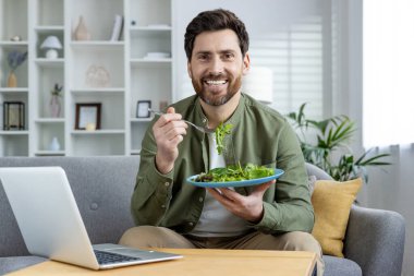 Evdeki kanepede oturan gülümseyen genç bir adamın portresi elinde bir defter, elinde sebzeli bir tabak ve taze salata ile kameraya güvenle bakıyor..