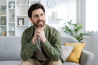 Gülümseyen genç bir adam evdeki kanepede oturuyor, ellerini yüzüne yakın tutuyor ve zevkle yan tarafa bakıyor..