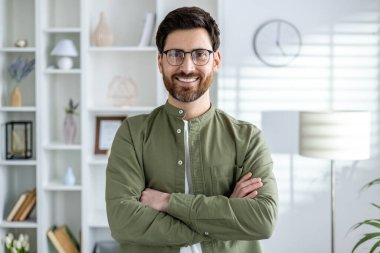 Yeşil tişörtlü ve gözlüklü bir gencin portresi bir ofiste duruyor, kollarını göğsünün üzerine koyuyor ve gülümseyerek kameraya bakıyor..