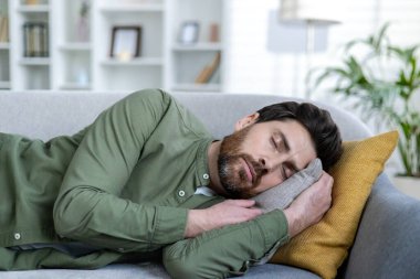 Kanepede uyuyan ve dinlenen genç bir adamın yakın plan fotoğrafı, gözleri kapalı uzanmış, elinde bir yastıkla..