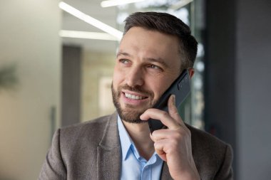 Modern bir ofis ortamında akıllı telefonuyla konuşan kendine güvenen bir iş adamı sohbet ederken gülümsüyor..