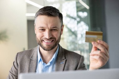 Modern bir ofis ortamında online alışveriş yaparken elinde kredi kartı tutan gülümseyen bir işadamı. E-ticaret ve finans kavramı.