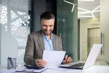 Gülümseyen işadamı belgeleri inceliyor ve modern ofis ortamında bir dizüstü bilgisayar üzerinde çalışıyor. Olumlu ve başarılı sonuçlara sahip profesyonel, üretken bir ortam.