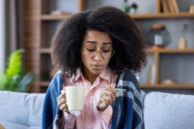Kadın kendini iyi hissetmiyor, battaniyeye sarılmış, elinde bir bardak tutuyor ve termometreyle ateşini ölçüyor..