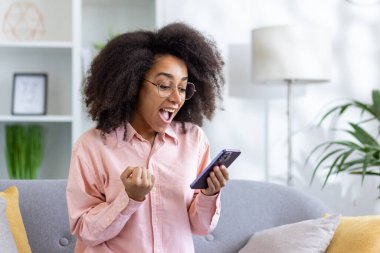 Pembe tişörtlü mutlu kadın telefonuna bakıyor, harika haberleri kutlarken evde bir kanepede oturuyor..