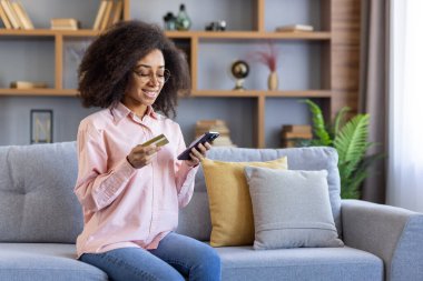 Gülümseyen kadın kredi kartını ve akıllı telefonunu online alışveriş için kullanıyor oturma odasında rahat bir kanepede oturuyor.