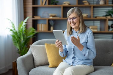Son sınıf öğrencisi bir kadın evinde bir kanepede otururken tablet kullanarak video görüşmesi yapıyor, gülümsüyor ve anın tadını çıkarıyor..