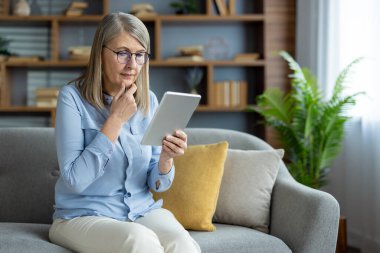 Kanepede oturan yaşlı bir kadın internette gezinmek ya da evde internet okumak için tablet kullanıyor.