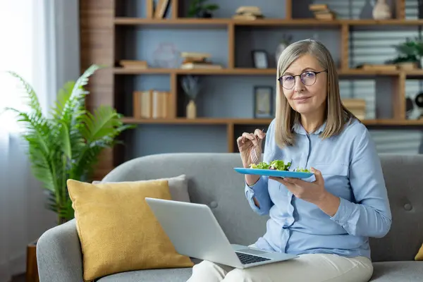 stock image Senior woman sitting on a couch, eating a healthy salad, and working on a laptop. Comfortable home setting with bookshelf and plants in the background.