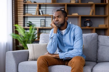 Evde dinlenen bir adam, düşünceli ve düşünceli bir ifadeyle bir kanepede oturuyor, kitaplıklar ve bitkilerle çevrili.
