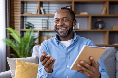 Elinde kalem ve defter tutan mutlu bir adam, çevrimiçi bir video görüşmesi sırasında gülümseyen ve konuşan, rahat bir oturma odasında oturan.
